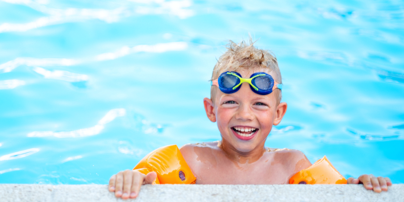 A young boy smiling at the edge of a pool and wearing swim goggles and floaties.